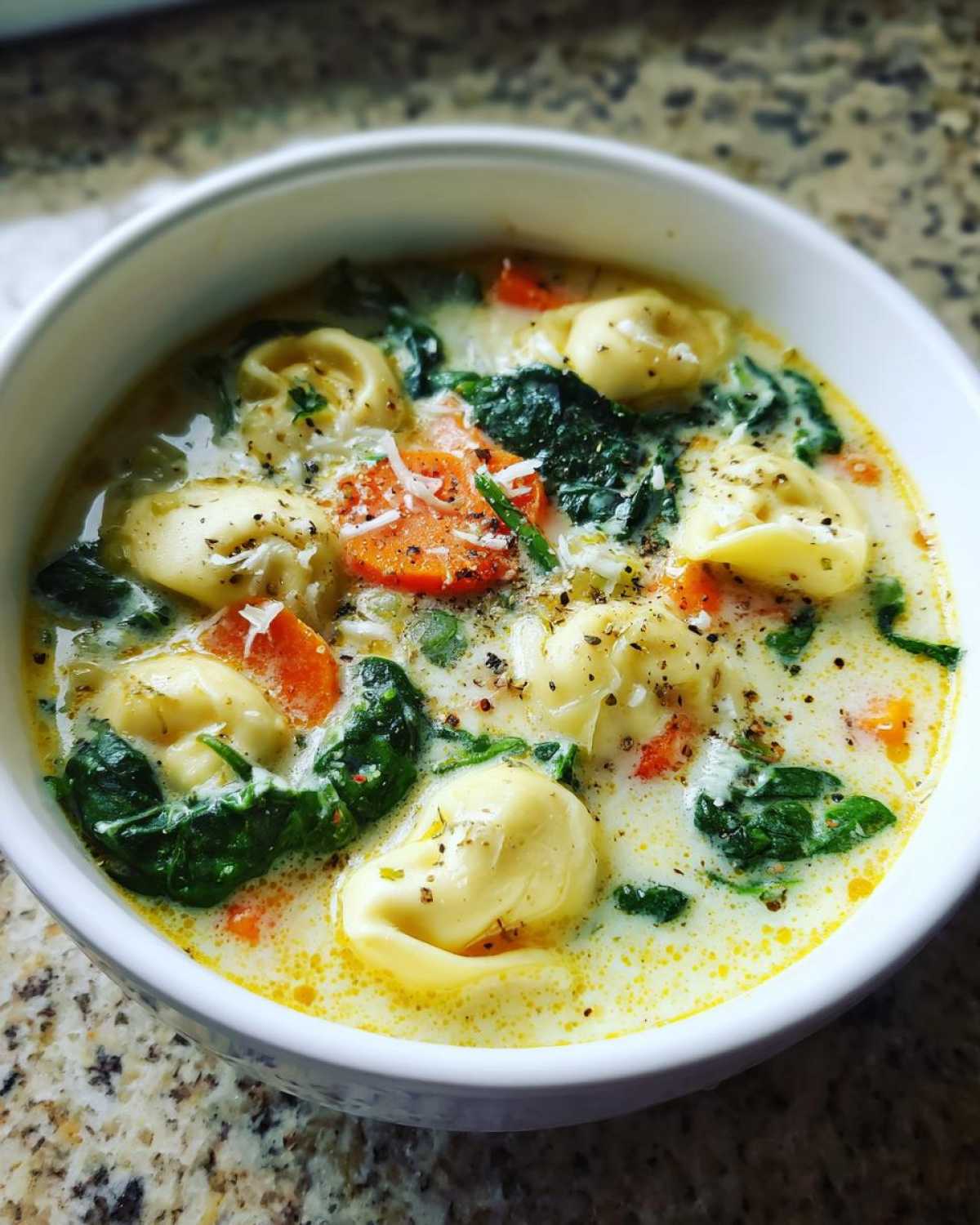 Close-up of a white bowl filled with One-Pot Creamy Tortellini Minestrone soup, featuring tortellini, spinach, and carrots.