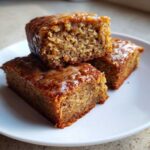 Three moist, square portions of Ooey Gooey Banana Bars stacked on a white plate, topped with a light glaze.