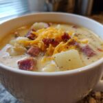Close-up of a creamy Pioneer Woman Sausage Potato Chowder topped with shredded cheddar cheese and bacon bits in a white mug.