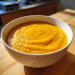 A close-up of a white bowl filled with vibrant orange Roasted Butternut Squash Soup, sitting on a wooden surface.