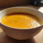 A close-up of a white bowl filled with vibrant orange Roasted Butternut Squash Soup, lightly seasoned with pepper.