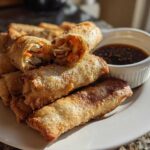 A plate of golden brown Sesame Veggie Spring Rolls, one cut open showing shredded vegetables inside, served with a side of dark dipping sauce.