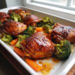 Close-up of glazed Sheet Pan Ginger Orange Chicken Thighs served with roasted broccoli and carrots in a white dish.