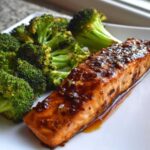 Close-up of a glazed Sheet Pan Honey Harissa Salmon fillet next to roasted broccoli florets on a white plate.