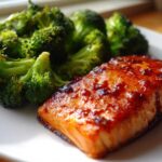 A close-up of a glazed Sheet Pan Honey Harissa Salmon fillet served next to bright green roasted broccoli florets on a white plate.