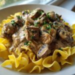 A close-up of a bowl featuring Slow Cooker Beef Stroganoff served over bright yellow egg noodles, garnished with parsley.