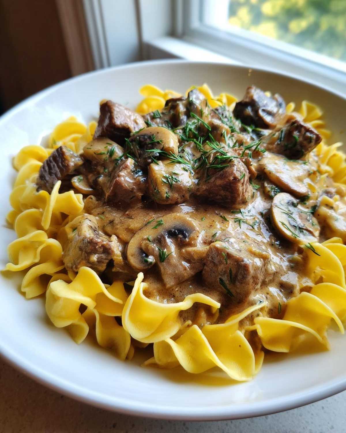 A close-up of a bowl of Slow Cooker Beef Stroganoff featuring tender beef chunks and mushrooms in a creamy sauce, served over wide egg noodles and garnished with dill.