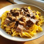 A close-up of a bowl filled with creamy Slow Cooker Beef Stroganoff served over bright yellow egg noodles.