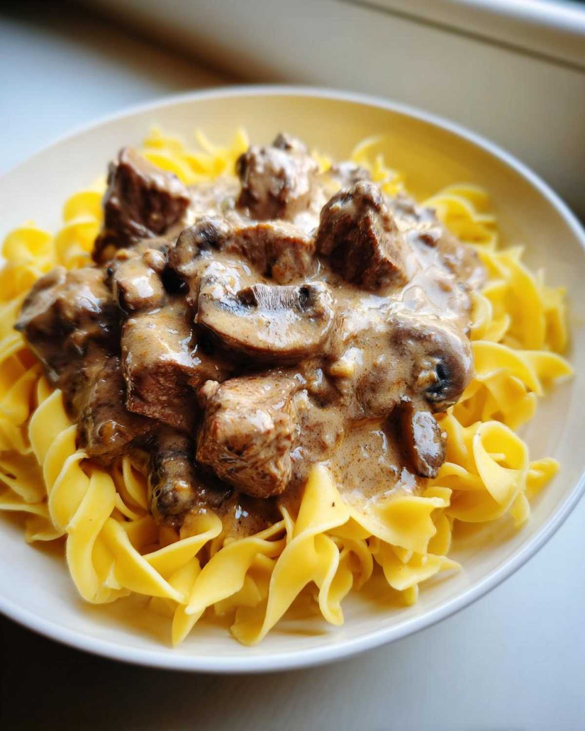 A close-up of a bowl filled with Slow Cooker Beef Stroganoff, featuring tender beef chunks and mushrooms in a creamy sauce over egg noodles.
