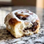 A close-up shot of a single, soft cinnamon roll with a bite taken out, showing the gooey filling and thick white frosting.