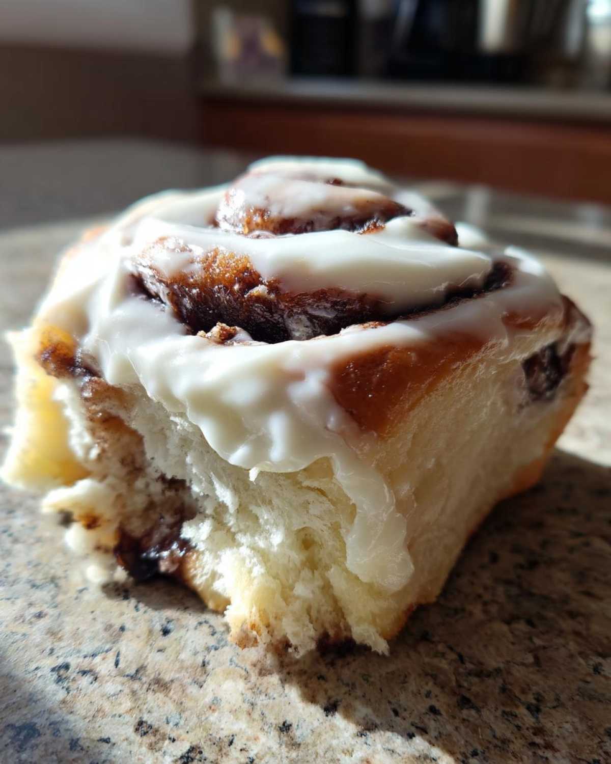 A close-up, sunlit photo showing a bite taken out of a soft Cinnamon Roll topped with thick white icing.