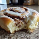 A close-up of one soft, fluffy cinnamon roll topped with thick white icing, showing a bite taken out.