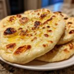 A stack of freshly made, soft Naan Bread brushed with melted butter, showing charred bubbles.