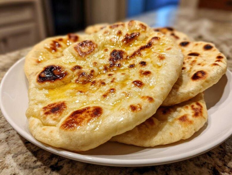 A stack of freshly made, soft Naan Bread brushed with melted butter, showing charred bubbles.