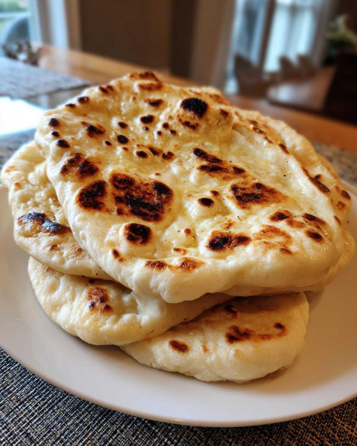 A stack of three freshly made, soft Naan Bread pieces resting on a white plate, showing golden-brown bubbles and char marks.