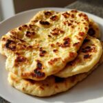 Close-up of three pieces of soft, golden Naan Bread stacked on a white plate, showing signature charred bubbles.