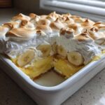 A square white dish filled with 8-layer Banana Pudding, showing a slice removed to reveal layers of custard, bananas, and topped with toasted meringue.