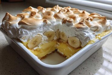 A square white dish filled with 8-layer Banana Pudding, showing a slice removed to reveal layers of custard, bananas, and topped with toasted meringue.