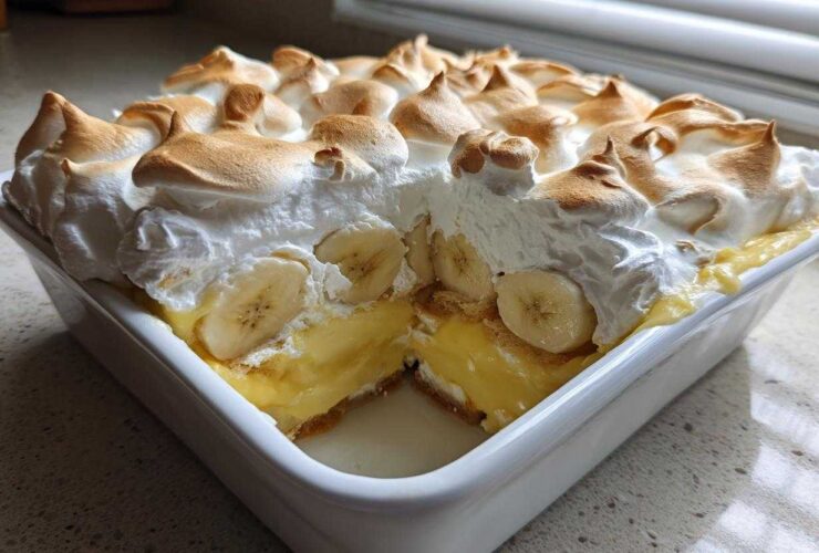 A square white dish filled with 8-layer Banana Pudding, showing a slice removed to reveal layers of custard, bananas, and topped with toasted meringue.