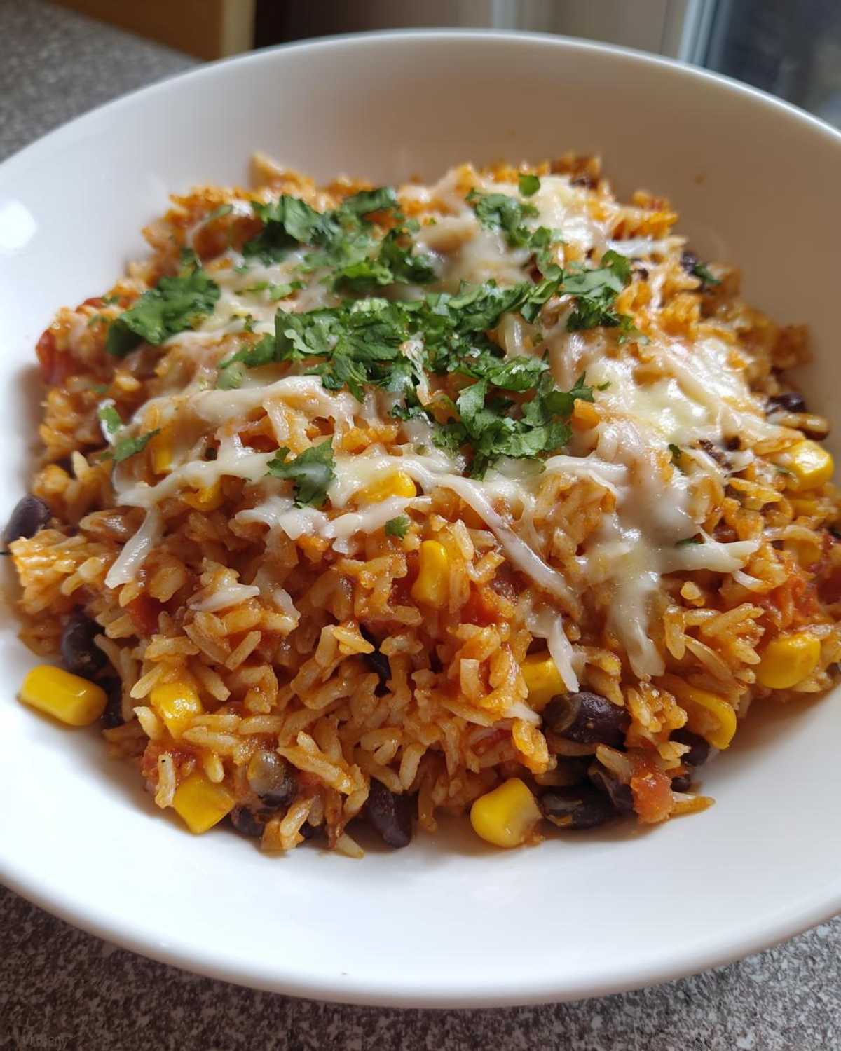 A close-up of a white bowl filled with Tasty Salsa Rice, black beans, corn, topped with melted white cheese and fresh cilantro.