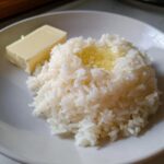 A serving of fluffy white Texas Roadhouse Seasoned Rice with melted butter pooled on top, next to a stick of butter.