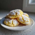 A pile of bright yellow Ultimate Lemon Cake Mix Cookies dusted heavily with white powdered sugar on a white plate.