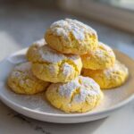 A stack of bright yellow Ultimate Lemon Cake Mix Cookies dusted generously with powdered sugar on a white plate.