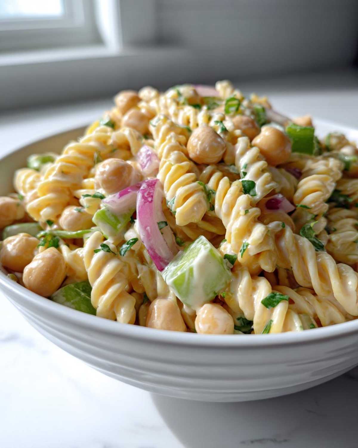 Close-up of Vegan Creamy Lemon Chickpea Pasta Salad featuring fusilli pasta, chickpeas, red onion, and green celery in a white bowl.