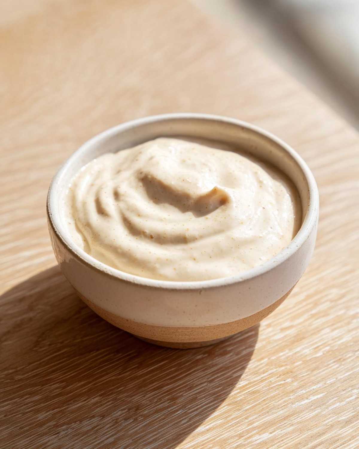 A small ceramic bowl filled with thick, creamy Vegan Creamy Lemon Tahini Dressing, sitting on a light wood surface.