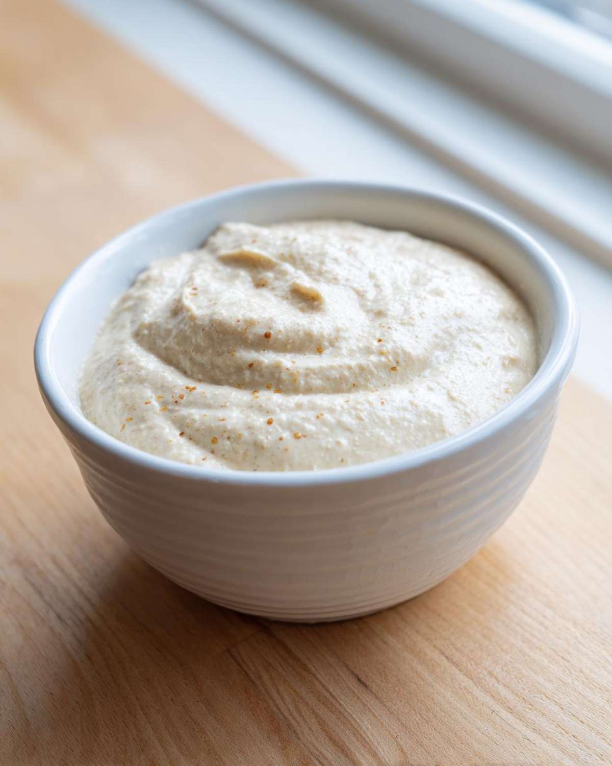 A white bowl filled with thick, creamy Vegan Creamy Lemon Tahini Dressing, resting on a light wooden surface.