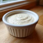 A close-up of thick Vegan Creamy Lemon Tahini Dressing swirled in a small white ceramic ramekin.