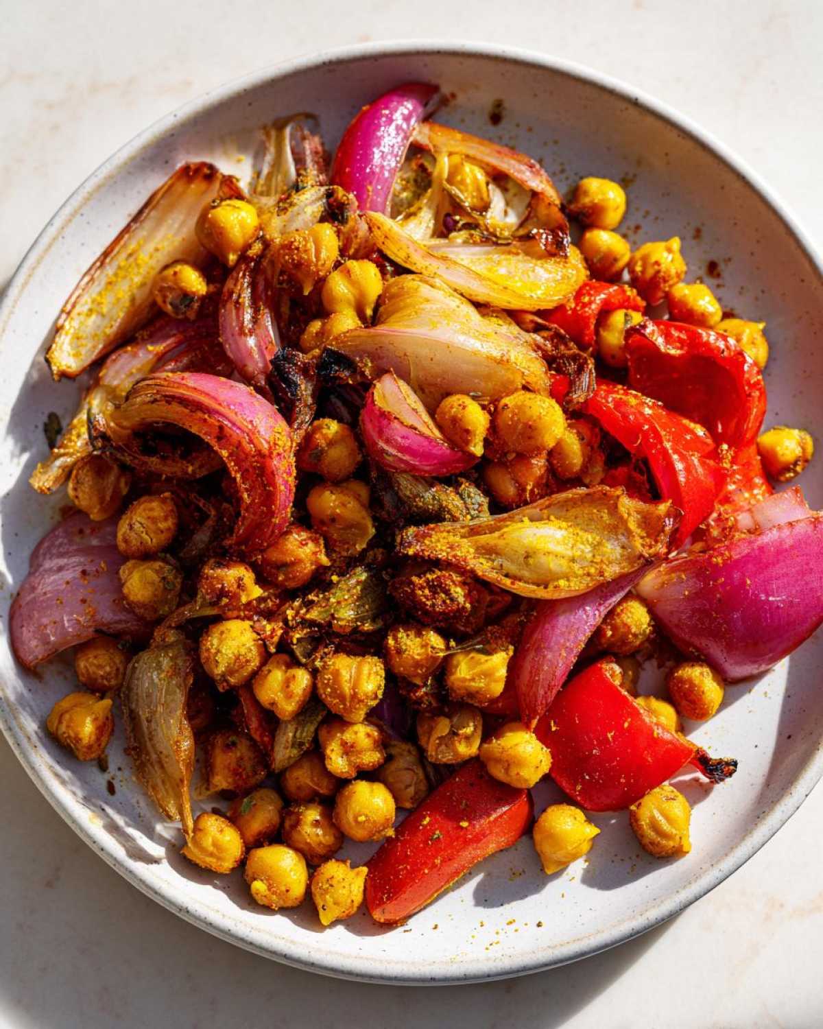Close-up of roasted chickpeas, red onion wedges, and red bell pepper pieces seasoned for Vegan One-Pan Chickpea Shawarma Sheet Bake.